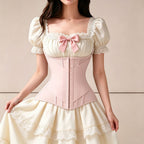 A woman wearing a cream-colored Victorian-style corset dress with a pale pink waist corset, puffed short sleeves with lace trim, a pink bow at the neckline, and a flared skirt with lace ruffles.