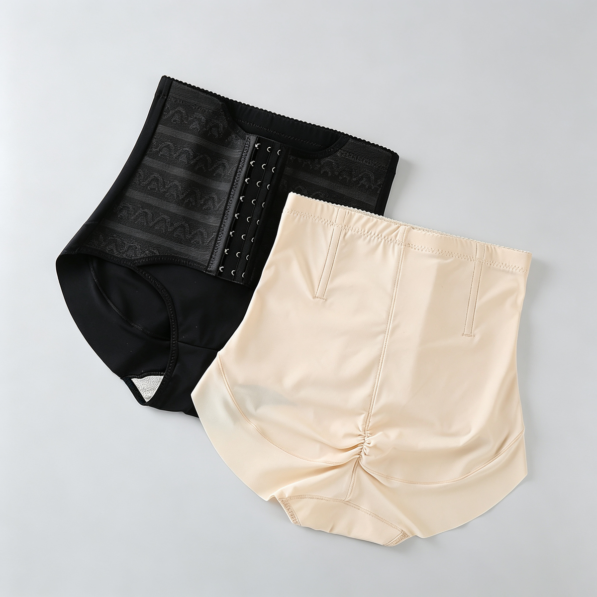 Two high-waisted compression briefs laid flat on a light gray background: one black with a lace-up front panel and one nude beige with a gathered front seam.