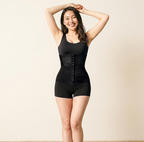A woman modeling a black waist trainer shapewear bodysuit with built-in shorts, standing against a plain beige background.