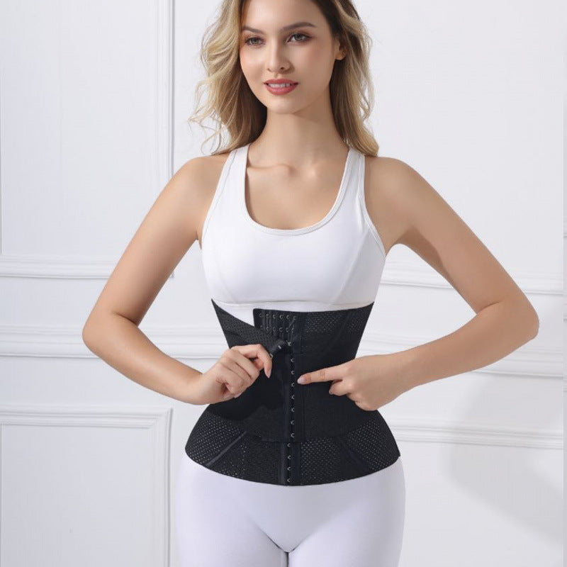 A woman wearing a white tank top and white leggings models a black waist trainer belt with a front zipper and lace-up closure against a white paneled wall.
