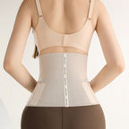 Rear view of a woman wearing a beige waist trainer shapewear belt over a matching beige bra and brown leggings, against a plain light background.