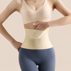 A woman modeling a beige waist trainer shapewear belt worn over a matching beige bra and blue leggings, set against a solid beige studio background.