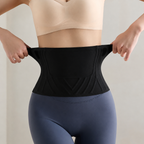 A person wearing a black waist trainer belt, stretching it with both hands to show its elasticity. The person is wearing a beige bra and slate blue leggings against a neutral beige background.