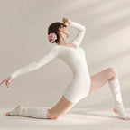 A woman in a white ribbed long-sleeve bodysuit and matching leg warmers poses dynamically on a light beige studio background, wearing a pink rose hair accessory.