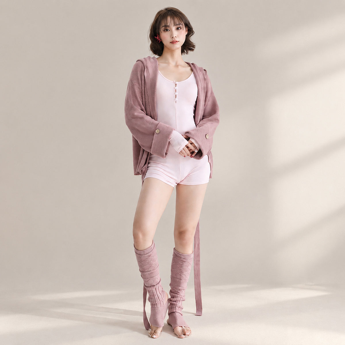 A woman models a coordinated lounge set consisting of a light pink bodysuit, a dusty mauve zip-up hoodie, matching mauve knee-high socks, and nude flat sandals against a warm beige studio backdrop with soft window shadows.