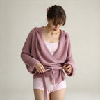 A woman wearing a dusty pink ribbed knit hooded wrap cardigan tied at the waist over a light pink tank top and matching pink shorts, photographed against a plain light beige studio background.