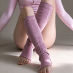 A person kneeling and crossing their legs while wearing pink knit leg warmers with textured diamond and cable-knit patterns, paired with a matching pink bodysuit.