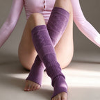 A model wearing purple cable-knit leg warmers with ribbed cuffs and open toes, paired with a light pink bodysuit, kneeling against a neutral beige background.
