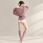 A woman stands barefoot in a studio wearing a dusty mauve oversized hoodie with a front knot detail, light pink shorts, and matching mauve fingerless leg warmers. She has a pink flower hair accessory in her hair.