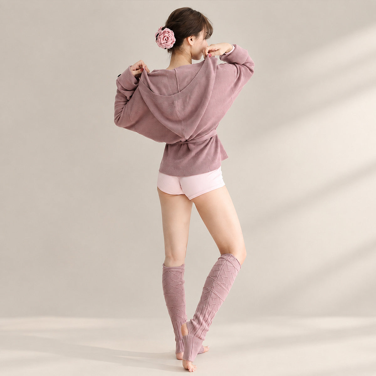 A woman stands barefoot in a studio wearing a dusty mauve oversized hoodie with a front knot detail, light pink shorts, and matching mauve fingerless leg warmers. She has a pink flower hair accessory in her hair.