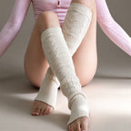 A person wearing cream-colored, cable-knit leg warmers with diamond-patterned knit sections, paired with a pink long-sleeve top, sitting against a neutral beige background.