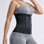 A woman wearing a black waist trainer belt with a front zip closure and a side pocket, paired with gray athletic wear, shown against a light gray studio background.