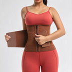 A woman wearing a brown waist trainer corset with a front zipper, holding the side panel of the device to demonstrate its flexibility. She is wearing a coral-red sports bra and matching leggings against a light gray studio background.