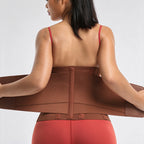 A woman shown from the back wearing a brown waist trainer belt over a red sports bra and red leggings, holding the sides of the belt outwards against a light gray studio background.