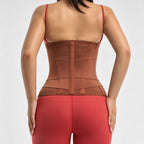 Rear view of a woman wearing a brown waist trainer corset with a center back zipper, paired with red leggings, against a light gray background.