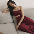 A woman reclining on a light gray sofa wearing a strapless deep burgundy two-piece set with a cutout waist and flowing pants.
