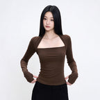 A woman modeling a fitted, long-sleeve brown top with a square neckline and a unique split neckline design, paired with black pants against a plain white studio background.