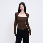 A woman modeling a fitted long-sleeve brown top with a square neckline and black pants, standing against a plain light gray studio background.
