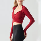 A woman modeling a long-sleeve red wrap-style athletic top with a deep plunging neckline, paired with black leggings, against a clean light gray studio background.