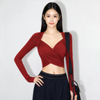 A woman modeling a long-sleeve wrap-style top in deep burgundy red, paired with dark navy pants and a black shoulder bag, against a plain light gray background.