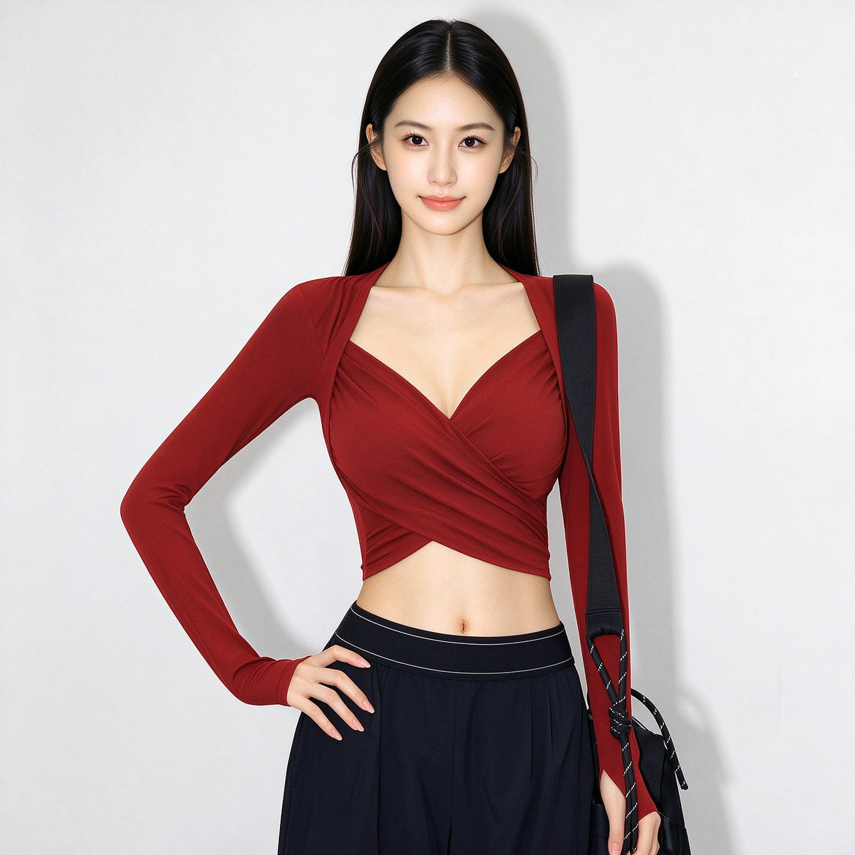 A woman modeling a long-sleeve wrap-style top in deep burgundy red, paired with dark navy pants and a black shoulder bag, against a plain light gray background.