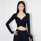 A woman modeling a black long-sleeve wrap-style crop top with a deep V-neckline, paired with a matching black skirt, against a plain light gray studio background.