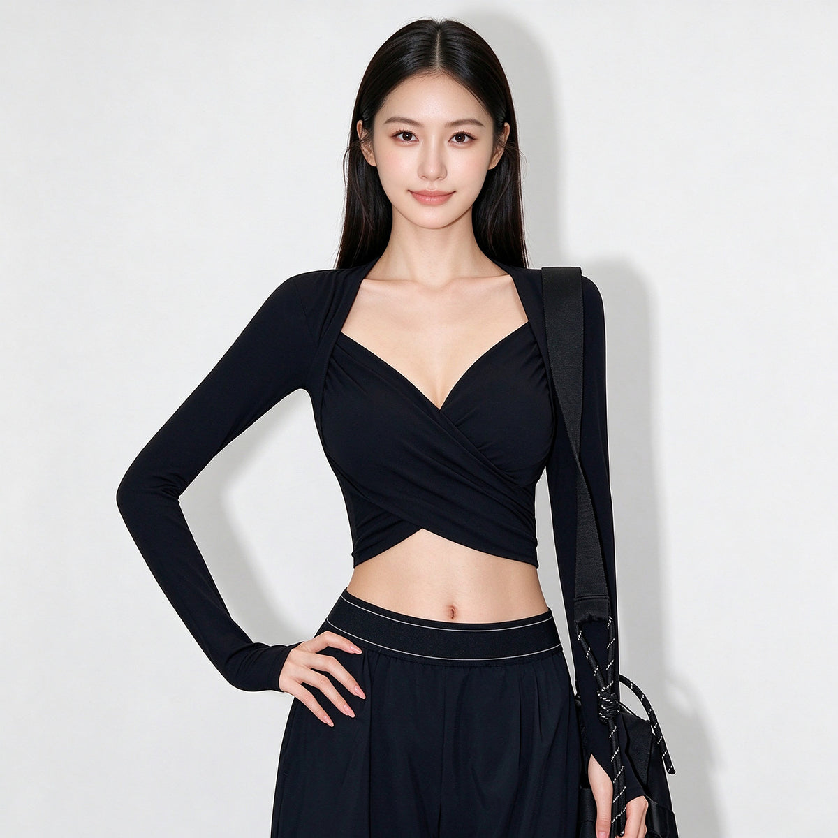 A woman modeling a black long-sleeve wrap-style crop top with a deep V-neckline, paired with a matching black skirt, against a plain light gray studio background.