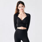 A woman modeling a black long-sleeve wrap-style crop top and matching black leggings against a plain light gray studio background.