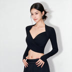 A woman modeling a black long-sleeve wrap-style crop top with a deep V-neckline, paired with a black skirt, set against a plain light gray studio background.