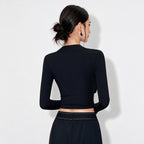 A woman shown from the back wearing a fitted black long-sleeve mock-neck crop top and black bottoms against a plain light gray studio background.