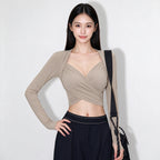A woman modeling a beige long-sleeve wrap-style crop top with a crossover V-neckline, paired with dark navy pants and a black shoulder bag, against a plain light gray background.