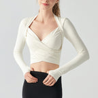 A woman wearing a fitted long-sleeve wrap-style crop top in warm off-white, paired with black bottoms, against a plain light gray studio background.