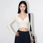A woman modeling a fitted long-sleeve wrap-style crop top in warm off-white, paired with dark navy pants and a black shoulder bag, against a plain light background.
