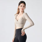 A woman modeling a beige long-sleeve wrap-style sports top with a crossover front and deep V-neckline, paired with black leggings, against a clean light gray studio background.