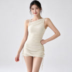 A woman modeling a beige one-shoulder ruched mini dress with side drawstrings against a plain light gray studio background.