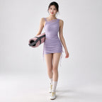 A woman stands against a light gray studio background wearing a lavender one-shoulder ruched mini dress and holding a rolled pink yoga mat with a dark strap.