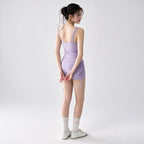 A woman stands in profile wearing a fitted lavender ruched mini dress with thin shoulder straps and side drawstrings, paired with white knee-high socks and white lace-up shoes, against a plain light gray studio background.